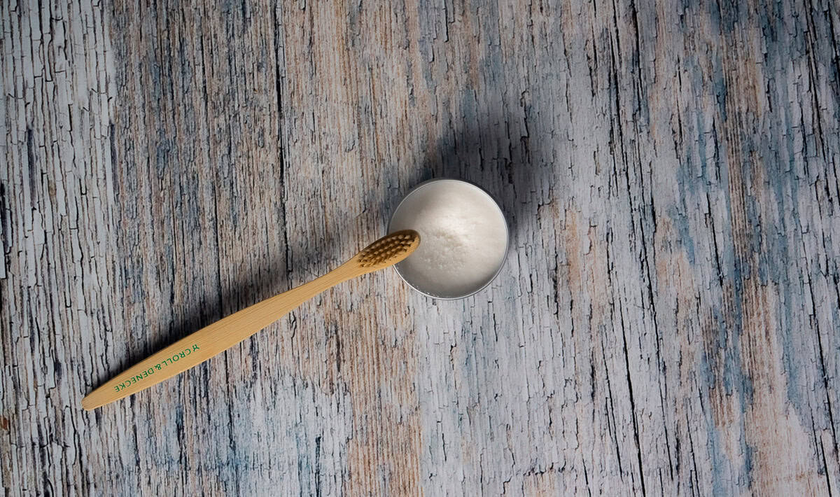 En bambustannbørste hviler på en liten boks fylt med hjemmelaget tannkrem, på en strukturert overflate av tre.