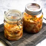 To glass med hjemmelaget kimchi med synlige grønnsaker er plassert på et grått klede mot en lys bakgrunn.