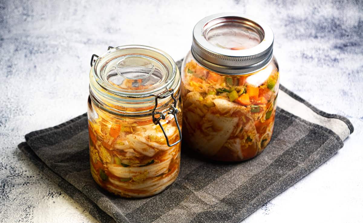 To glass med hjemmelaget kimchi med synlige grønnsaker er plassert på et grått klede mot en lys bakgrunn.