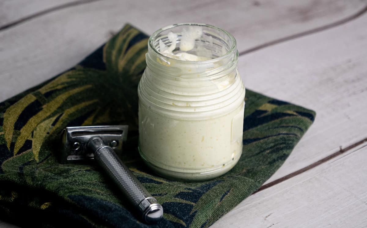 En glasskrukke med barberkrem står på et sammenbrettet, grønnmønstret tøystykke ved siden av en barberhøvel i metall på en lys treflate.