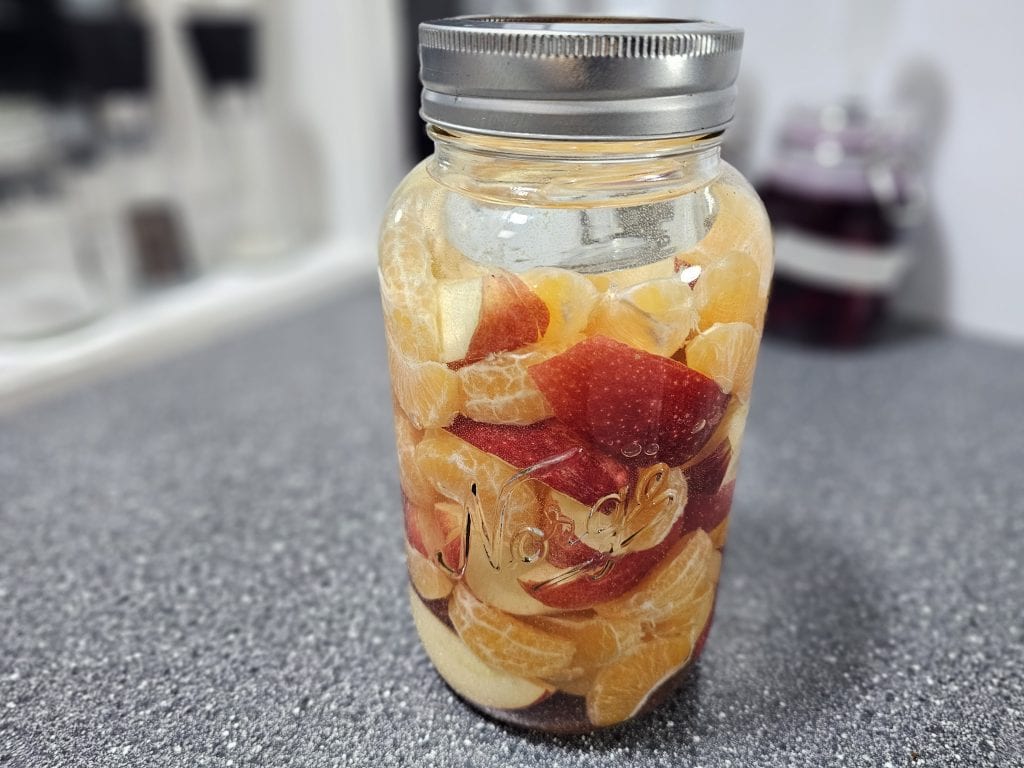 På en grå benkeplate står en glasskrukke fylt med skrelte appelsinbiter og røde epleskiver.