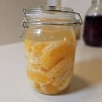 En glasskrukke fylt med skrelte appelsinbiter i væske står på en hvit flate, med en annen krukke uskarpt i bakgrunnen. Fermenterte appelsiner.