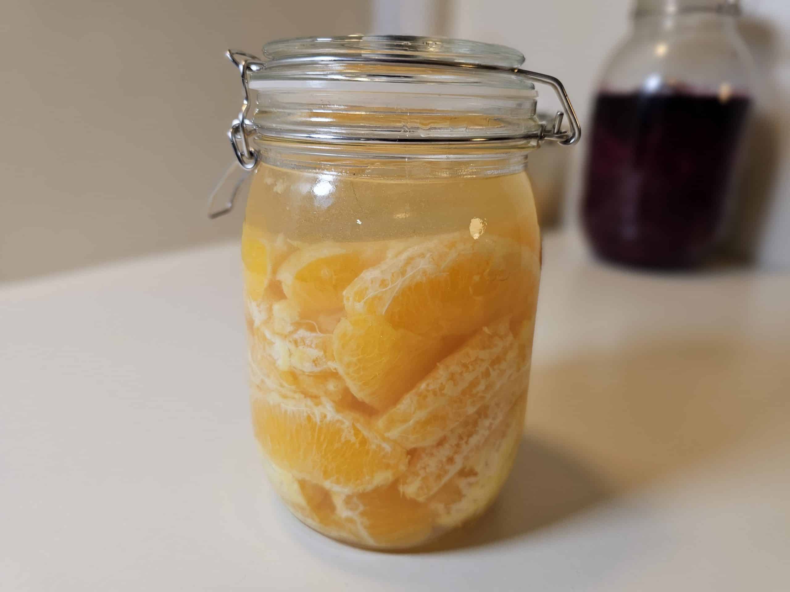 En glasskrukke fylt med skrelte appelsinbiter i væske står på en hvit flate, med en annen krukke uskarpt i bakgrunnen. Fermenterte appelsiner.