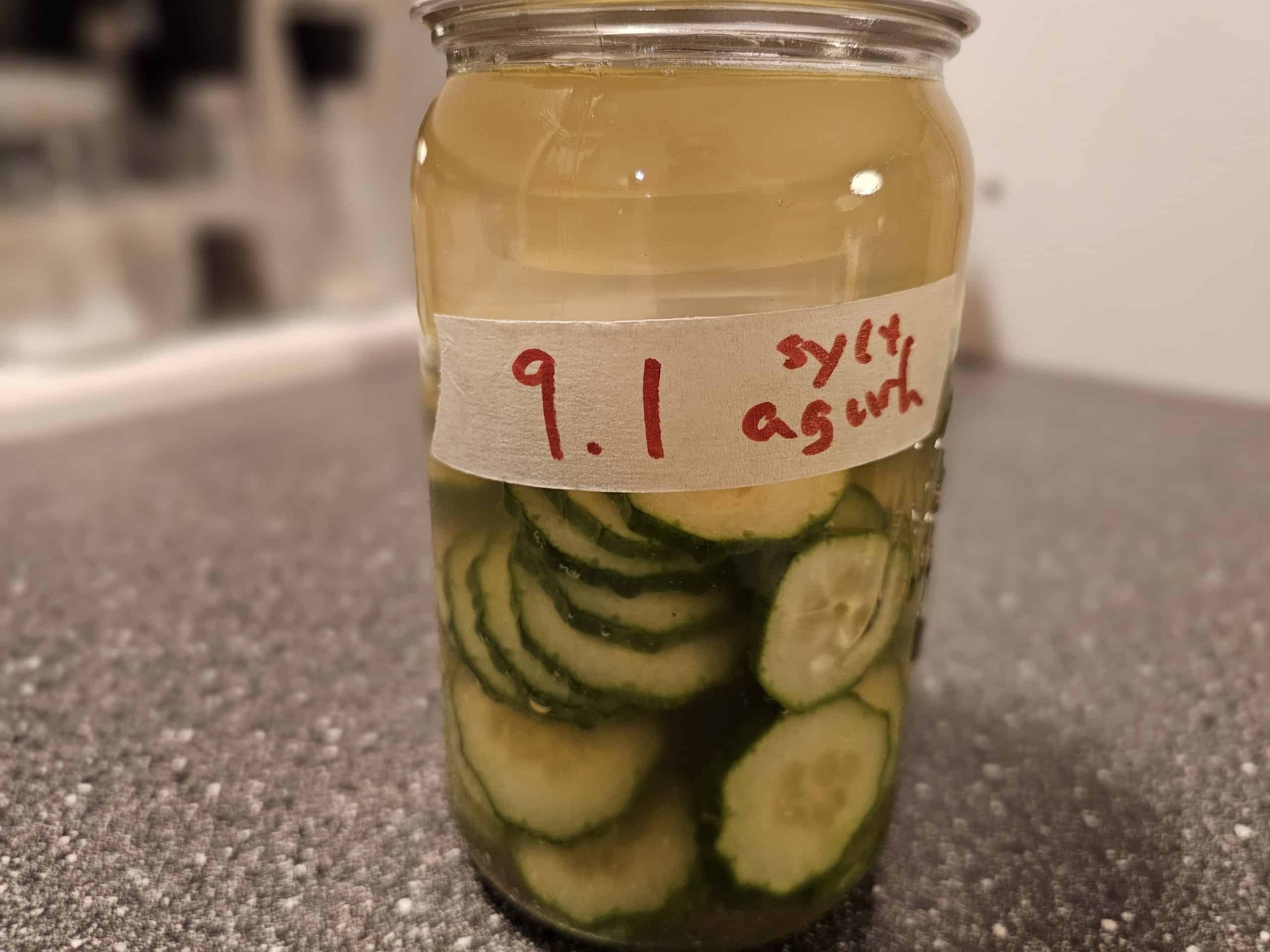 På en mørk benkeplate står en glasskrukke fylt med agurkskiver og væske, merket "9.1 sylt agurk" med rød tusj. Sylteagurk.