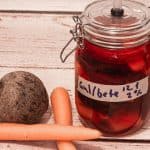 Et glass med fermenterte gulrøtter og beter, merket "gul/bete 2 %" står på en treflate ved siden av en rødbete og to gulrøtter.