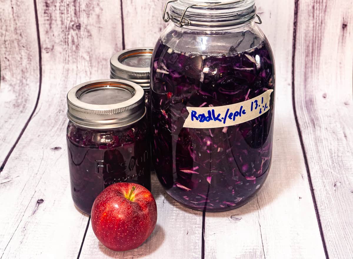 Tre glasskrukker fylt med fermentert rødkål står utstilt på en rustikk treflate sammen med et rødt eple.