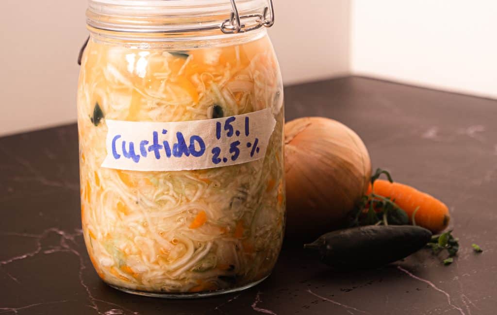 Et glass merket "Curtido" fylt med fermenterte grønnsaker er plassert på en mørk flate med en løk, gulrot og jalapeño ved siden av.