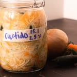 Et glass merket "Curtido" fylt med fermenterte grønnsaker er plassert på en mørk flate med en løk, gulrot og jalapeño ved siden av.