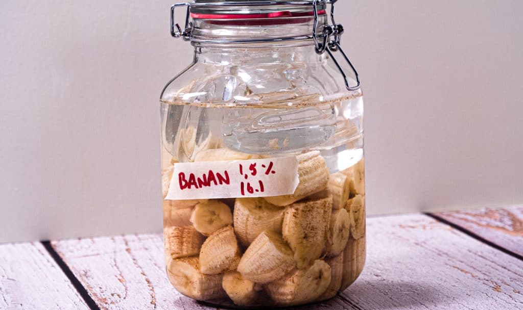 En glasskrukke fylt med fermenterte bananskiver og væske, plassert på en treflate mot en lys bakgrunn.