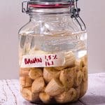 En glasskrukke fylt med fermenterte bananskiver og væske, plassert på en treflate mot en lys bakgrunn.