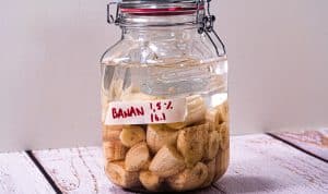 En glasskrukke fylt med fermenterte bananskiver og væske, plassert på en treflate mot en lys bakgrunn.