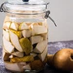 På en mørk benkeplate står en glasskrukke med fermenterte pæreskiver i væske ved siden av en hel pære.
