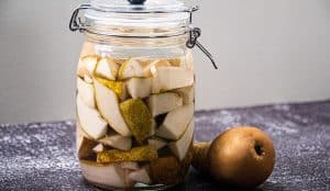 På en mørk benkeplate står en glasskrukke med fermenterte pæreskiver i væske ved siden av en hel pære.