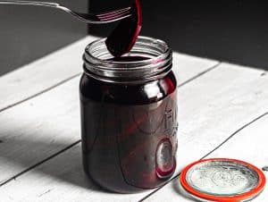 En gaffel holder en syltet rødbetskive over en åpen glasskrukke fylt med syltede rødbeter; metallokket hviler på den hvite treflaten ved siden av krukken.