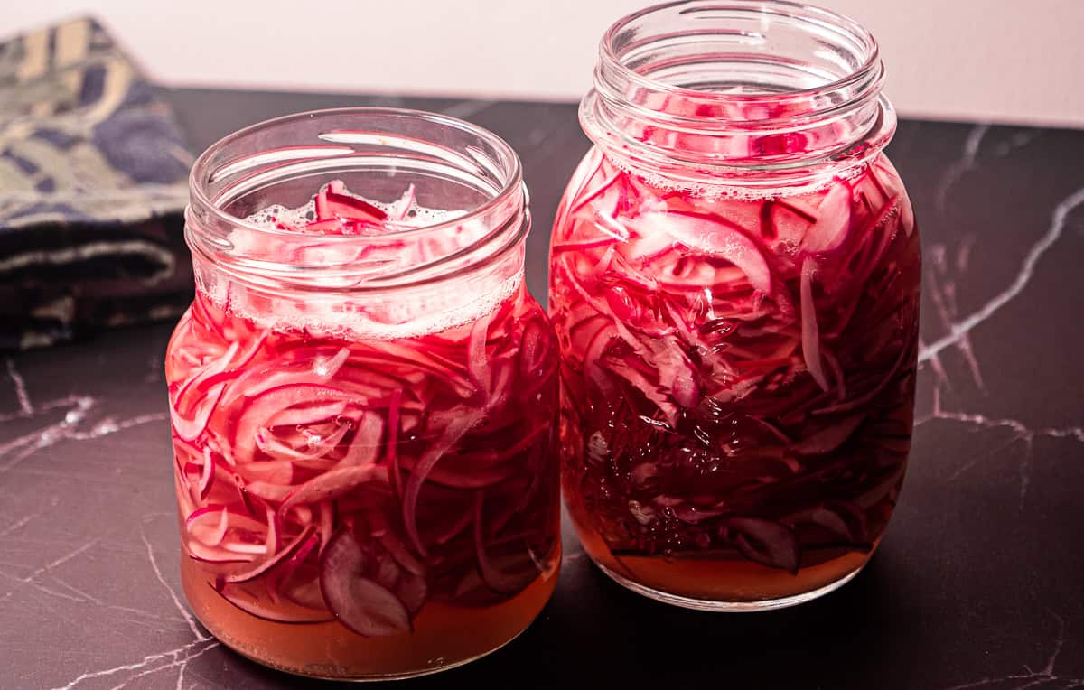 To glasskrukker fylt med tynne skiver syltet rødløk som ligger i en rosa væske, plassert på en mørk marmorplate.
