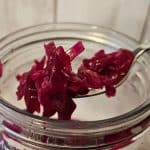 En gaffel løfter fermentert rødkål/sauerkraut fra en åpen glasskrukke mot en lys trebakgrunn.