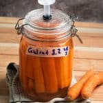 I et glass med gjæringslokk ligger gulrøtter i væske. Ved siden av glasset ligger to hele gulrøtter på et underlag av tre.