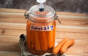 I et glass med gjæringslokk ligger gulrøtter i væske. Ved siden av glasset ligger to hele gulrøtter på et underlag av tre.