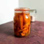 En glasskrukke fylt med syltede gulrøtter, står på en lilla flate med en uskarp beholder i bakgrunnen.