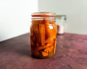 En glasskrukke fylt med syltede gulrøtter, står på en lilla flate med en uskarp beholder i bakgrunnen.