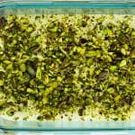 Et glassfat fylt med hjemmelaget pistasjis toppet med et tykt lag hakkede pistasjnøtter, med noen hele pistasjnøtter spredt på overflaten like ved.