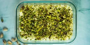 Et glassfat fylt med hjemmelaget pistasjis toppet med et tykt lag hakkede pistasjnøtter, med noen hele pistasjnøtter spredt på overflaten like ved.