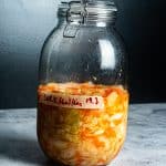 På en grå flate mot en mørk bakgrunn står en stor glasskrukke fylt med fermenterte grønnsaker, surkål, gulrøtter