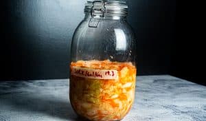 På en grå flate mot en mørk bakgrunn står en stor glasskrukke fylt med fermenterte grønnsaker, surkål, gulrøtter