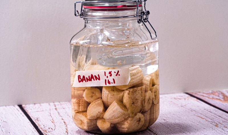En glasskrukke fylt med fermenterte bananskiver og væske, plassert på en treflate mot en lys bakgrunn.