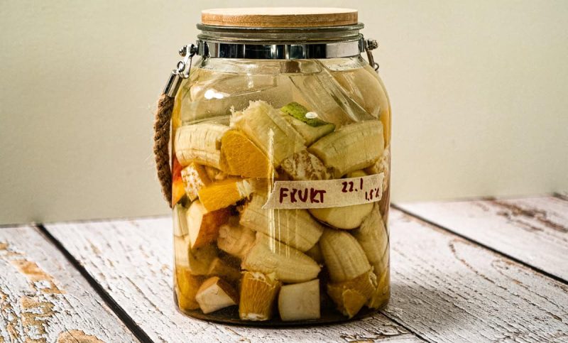 En glasskrukke med trelokk inneholder oppkuttede fermenterte fruktbiter merket "FRUKT 22,1", som står på en lys treflate.