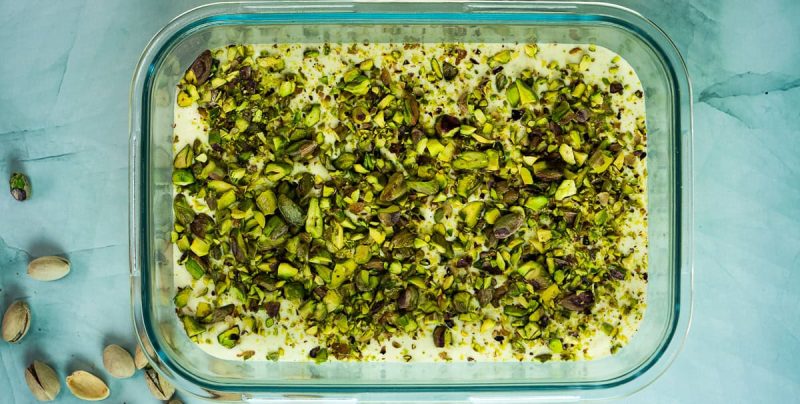 Et glassfat fylt med hjemmelaget pistasjis toppet med et tykt lag hakkede pistasjnøtter, med noen hele pistasjnøtter spredt på overflaten like ved.