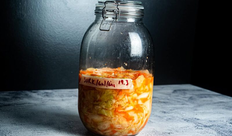 På en grå flate mot en mørk bakgrunn står en stor glasskrukke fylt med fermenterte grønnsaker, surkål, gulrøtter