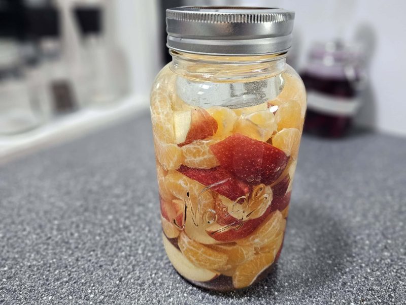 På en grå benkeplate står en glasskrukke fylt med skrelte appelsinbiter og røde epleskiver.